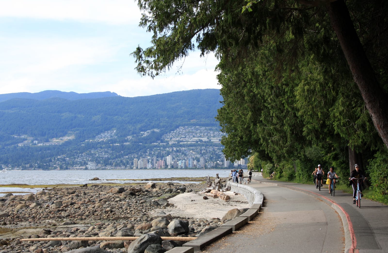 Cyclists on the Stanley Park Seawall beside a rocky shore, with the North Shore hills across the water